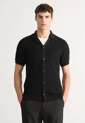 Black short-sleeve button-up shirt featuring a textured knit design, notched collar, and ribbed hem. Simple and modern construction.