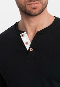 Camisa henley negra con cuello redondo, presenta una tapeta blanca y tres botones de madera. Textura suave, con mangas cortas visibles.