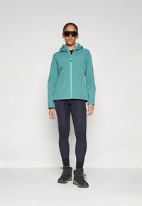 Casaco impermeável verde-azulado com capuz, fecho de correr frontal e bolsos laterais; combinado com leggings pretas e botas pretas robustas.