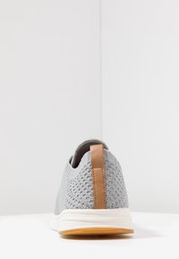 Sneaker grigia in maglia con superficie testurizzata, linguetta in pelle marrone sul tallone e intersuola bianca. Suola in gomma con un leggero motivo di aderenza.