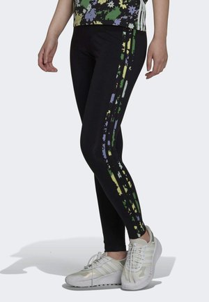 Černé legíny s texturovaným, vícerozměrným vzorem v zelené, žluté a fialové barvě po stranách, doplněné bílými sportovními botami.