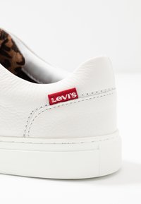 Baskets en cuir blanc avec une surface texturée, une étiquette Levi's rouge contrastante, et une semelle en caoutchouc avec un motif de marche discret. Intérieur avec imprimé léopard.