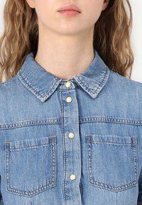 Chemise en denim avec un col classique, couleur bleu clair, fermeture à boutons sur le devant, deux poches poitrine et des détails de couture visibles.