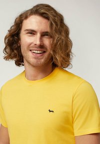 La t-shirt gialla in cotone presenta un collo rotondo e maniche corte, con un piccolo logo di un bassotto nero sul lato sinistro del petto. Tessuto morbido.