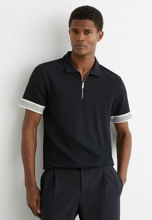 Férfi fekete rövid ujjú galléros pólóban, fehér csíkos mandzsettával és elöl cipzárral, egyik kezével zsebre tett kézzel áll fehér fal előtt.