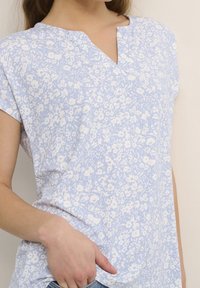Woman wearing a light blue floral short-sleeve top with a small V-neck, hand resting near the waist.