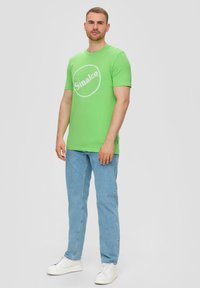 Groene katoenen T-shirt met een wit "Sinalco"-logo, gecombineerd met lichtblauwe jeans en witte sneakers. Eenvoudige ronde halslijn en korte mouwen.