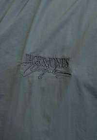 Jasnoszara tkanina o lekko teksturowanym wykończeniu, z haftowanym logo "JACK & JONES" w ciemnoszarym włóknie.