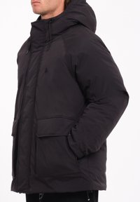 Zwarte pufferjack met een capuchon, voorzien van een rits- en knoopsluiting, twee zijzakken en een eenvoudig logo op de bovenste linkerborst.