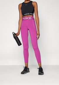 Leggings compressivi rosa con vita alta e logo grigio "NIKE PRO"; abbinati a una maglietta senza maniche nera e scarpe da atletica nere.