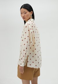 Cream long-sleeve shirt with red polka dots, buttoned collar, and a single chest pocket. Model wears brown shorts, displaying a relaxed fit.