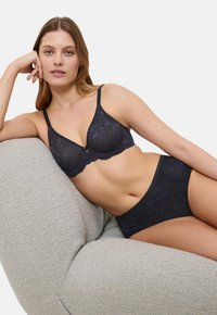 Žena se opušta na teksturiranoj fotelji noseći odgovarajući tamnoplavi čipkasti grudnjak i gaćice visokog struka.
