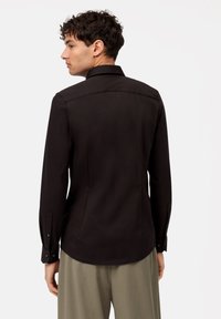 Camicia nera a maniche lunghe con colletto strutturato, caratterizzata da polsini con bottoni e una texture liscia e raffinata. Il design sul retro presenta un taglio sottile.