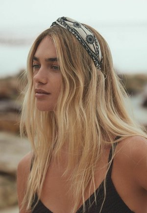 Junge Frau mit langen blonden Haaren, die ein gemustertes Stirnband und ein schwarzes Tanktop trägt, schaut im Freien nachdenklich zur Seite.