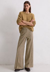 Vrouw staat met armen gekruist, gekleed in een mosterdkleurige trui, beige wijde geruite broek en donkerbruine schoenen tegen een plain witte muur.