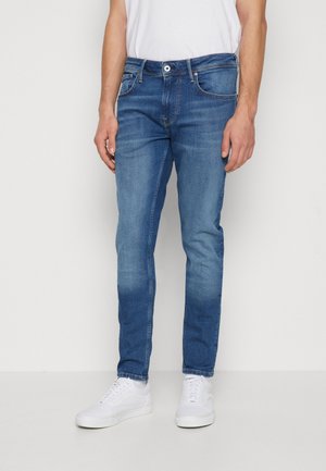 Man die een slim-fit blauwe jeans en witte sneakers draagt, staand tegen een effen witte achtergrond.