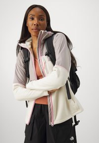 Φλις μπουφάν σε κρεμ και γκρι, με ροζ φερμουάρ, καπελάκι και λεπτομέρειες στους ώμους. Φορεμένο με σακίδιο, αναδεικνύει τη λειτουργική του σχεδίαση.