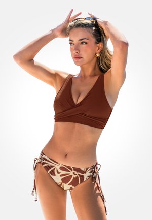 Kobieta poprawiająca okulary przeciwsłoneczne na głowie, ubrana w brązowy top bikini wiązany oraz kwiatowe majtki bikini z bokami wiązanymi na tle jednolitym.