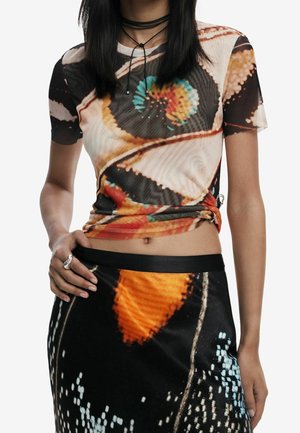 Mujer vistiendo un conjunto de top corto y falda con estampado de alas de mariposa a juego, con un collar de cordón negro en capas y un anillo plateado.