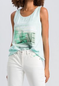 Mujer vistiendo una blusa sin mangas de color azul claro con texto verde y jeans de talle alto blancos, contra un fondo liso.
