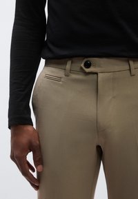 Μπεζ παντελόνι από λείο ύφασμα, με μπροστινό κουμπί, φερμουάρ και πλαϊνές τσέπες με κομψό σχεδιασμό. Ορατό μαύρο μπλουζάκι με μακριά μανίκια.