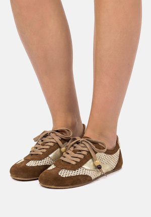 Zapatillas de ante marrón y beige con paneles de malla y cuentas decorativas en los cordones, usadas en pies descalzos sobre un fondo blanco.
