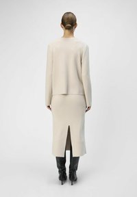 Pull beige et jupe ajustée, tissu texturé, détail d' fente à l'arrière, associé à des bottes noires jusqu'aux genoux. Design simple et épuré.