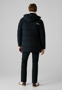 Chaqueta acolchada azul oscuro con capucha, que presenta un collar de borreguito, puños elásticos y un logo bordado en la parte superior de la espalda. Combinada con jeans oscuros.