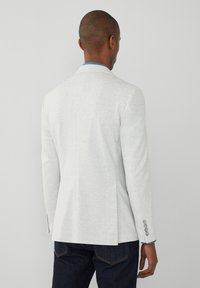 Helles grauer Blazer mit taillierter Passform, einer Schlitze hinten und dreiknöpfigen Details an den Ärmeln. Über einem blauen Hemd getragen.