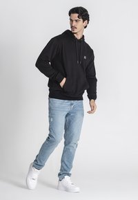 Felpa nera in cotone con tasca frontale, cappuccio con coulisse e logo piccolo. Abbinata a jeans chiari strappati e sneakers bianche.