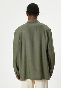 Mand iført en løstsiddende olivengrøn langærmet skjorte og lysebeige bukser, stående med ryggen til mod en ensfarvet hvid baggrund.