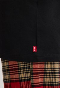 Zwart katoenen top met een rood label met een uitroepteken aan de zoom, gecombineerd met rode, zwarte en tan geruite broek.