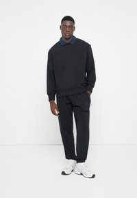 Felpa nera con colletto blu navy, abbinata a pantaloni neri, indossata con sneakers bianche e grigie. Design semplice e vestibilità rilassata.