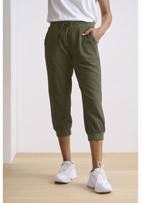 Olijfgroene capri-broeken van zachte stof, met zijzakken, een elastische tailleband met een trekkoord en opgerolde zoom. Paar witte sneakers.