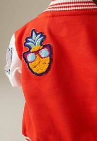 Gros plan du dos d'une veste rouge avec un écusson brodé d'un ananas portant des lunettes de soleil rouges et une manche blanche partiellement visible avec un autre écusson.