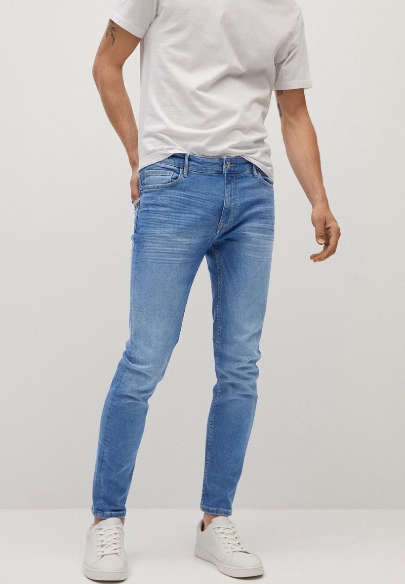 Vaqueros de denim azul claro con un corte slim, textura desgastada, diseño de cinco bolsillos y cierre de botón metálico, combinados con una camiseta blanca y zapatillas blancas.