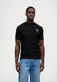 Μαύρο κοντομάνικο T-shirt από βαμβάκι, με μια μικρή σκούρα μπλε γραφική εκτύπωση στην αριστερή περιοχή του στήθους. Το μοντέλο συνδυάζεται με μπλε τζιν.