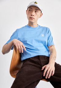Camiseta de algodón azul claro con cuello acanalado, que presenta un pequeño logo blanco. Pantalones de pana marrones, sentado en una silla de madera.