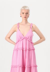 Pink tiered dress featuring ruffled straps, a deep V-neckline, and textured fabric. Designed with horizontal ruffles at each tier.