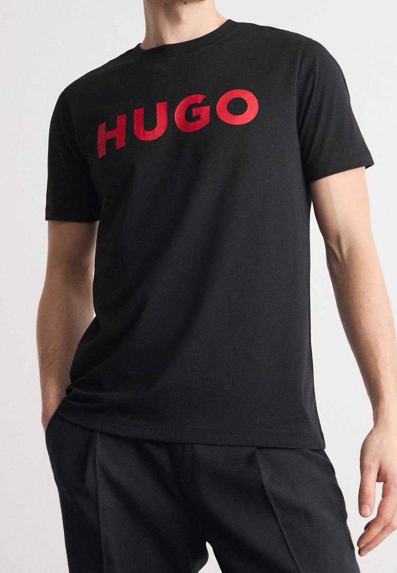 Schwarzes Baumwoll-T-Shirt mit einem großen roten "HUGO"-Logo auf der Brust. Kurze Ärmel und klassischer Rundhalsausschnitt. Glatte Textur.