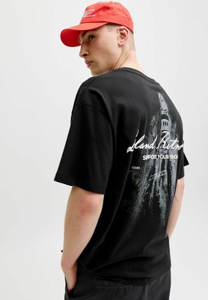 Jeune homme portant une casquette rouge et un T-shirt noir avec un texte blanc et un design graphique, posant de profil sur un fond clair uni.