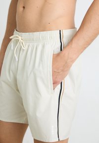Shorts de bain pour hommes beiges avec une taille élastique, fermeture à cordon de serrage, poches latérales et rayures noires et dorées sur les côtés. Marque sur la jambe.