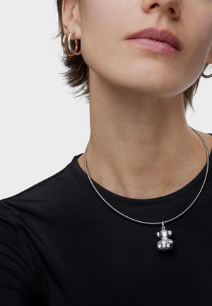 Colgante en forma de oso plateado sobre una delgada cadena metálica, usado sobre una camiseta negra ajustada. Se ve un pendiente de aro dorado en la oreja izquierda.