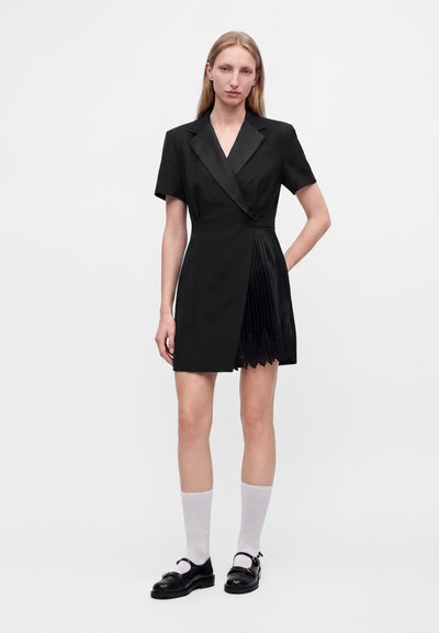 Jeune femme debout portant une robe blazer noire à manches courtes avec un détail plissé, des chaussettes blanches et des chaussures plates noires sur un fond uni.