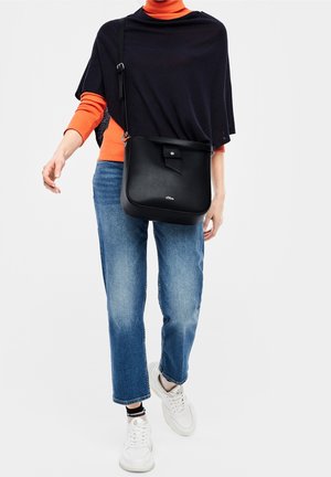 Osoba ubrana w pomarańczową bluzę z długim rękawem, czarny poncho, niebieskie dżinsy, białe trampki, niosąca czarną torebkę na ramię.