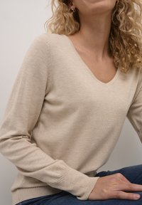 Pull en tricot beige avec un col en V, manches longues, poignets côtelés et une texture douce. Associé avec un jean foncé.