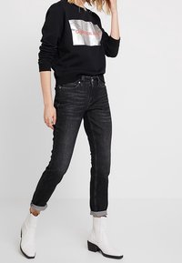 Felpa nera con logo metallizzato, abbinata a jeans in denim scuro e stivali bianchi alla caviglia. I jeans sono arrotolati alle caviglie.