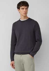 Ciemnoszary sweter z długim rękawem o prążkowanej fakturze i okrągłym dekoltem, zestawiony z jasnooliwkowymi spodniami. Tkanina wydaje się miękka i lekka.