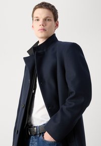 Cappotto di lana blu navy con colletto alto e chiusura con bottoni. Presenta tasche frontali angolate e cuciture sulle spalle. Indossato sopra una camicia bianca e jeans.