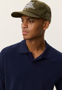 Καπέλο ελιάς πράσινο με γραφικό σχέδιο στο μπροστινό μέρος και λευκό κείμενο· μπλε ναυτικό πόλο πουκάμισο με γιακά και διακριτική κεντημένη λεπτομέρεια λογότυπου.